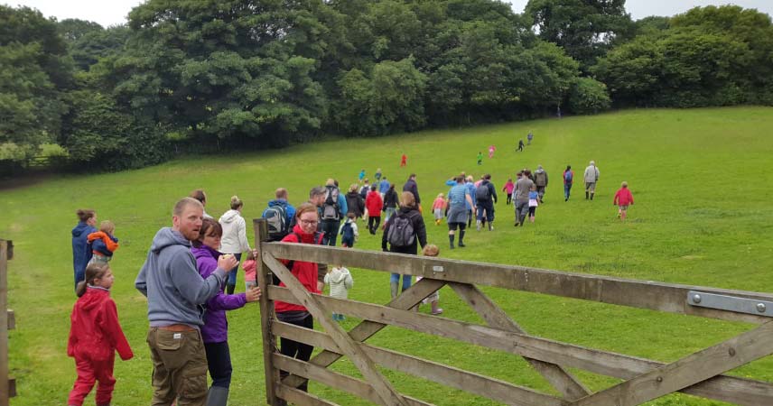 Large group of adults and children walking across a field as one person holds open a gate.