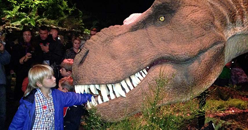 Boy with his hand in a model dinosaur's mouth at the Eden Project whilst surrounded by people.
