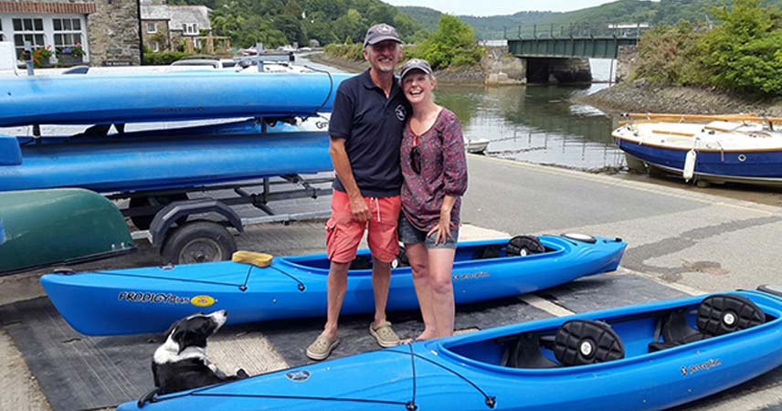 Two adults and their dog posing by a range of kayaks next to a river in Cornwall.