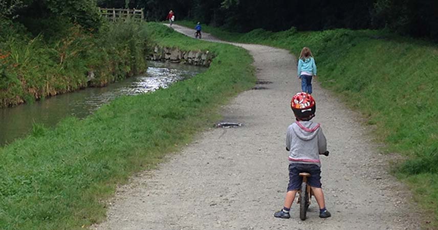 Child riding a bike by a river in Cornwall along a path, with two other children in front of him.