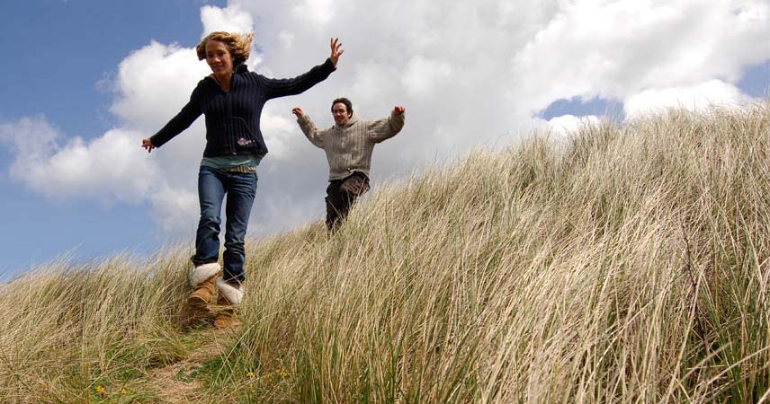 Two adults running through the sand dunes in Cornwall on a sunny day.