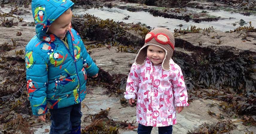 Child and toddler in coats and hats smiling for the camera, whilst on a beach covered in seaweed.