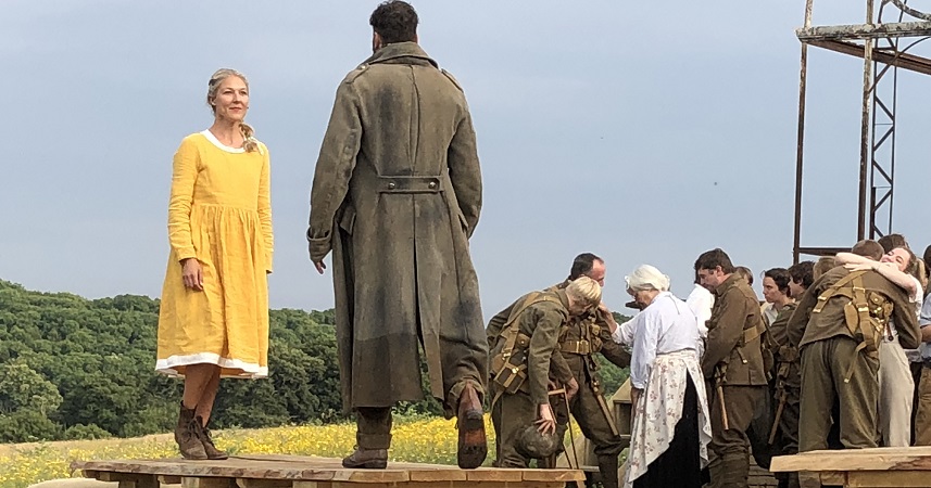 Two actors, one in yellow, the other in an army uniform, stood on a wooden bench table, in front of a group of more actors wearing army uniforms and hugging
