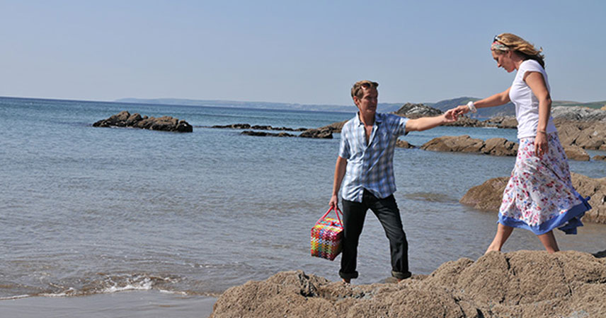 Man helping woman off some rocks while holding a picknick basket on the beach at high tide