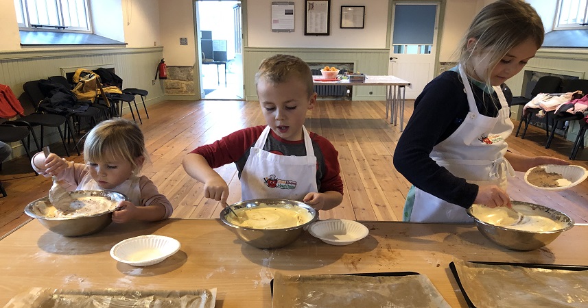 Three children wearing Little Kitchen Magicians aprons mixing ingredients in metal bowls.