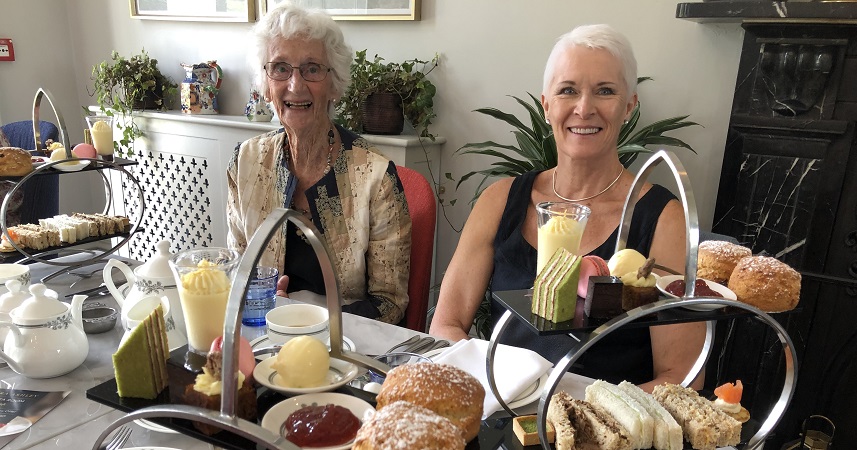Two well dressed ladies sat at a table with afternoon tea stands, including small cakes, scones and sandwiches