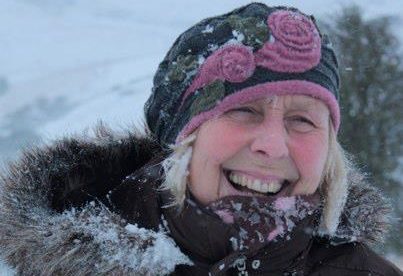 Pat in brown winter coat with faux fur collar and pink and blue woolly hat, laughing