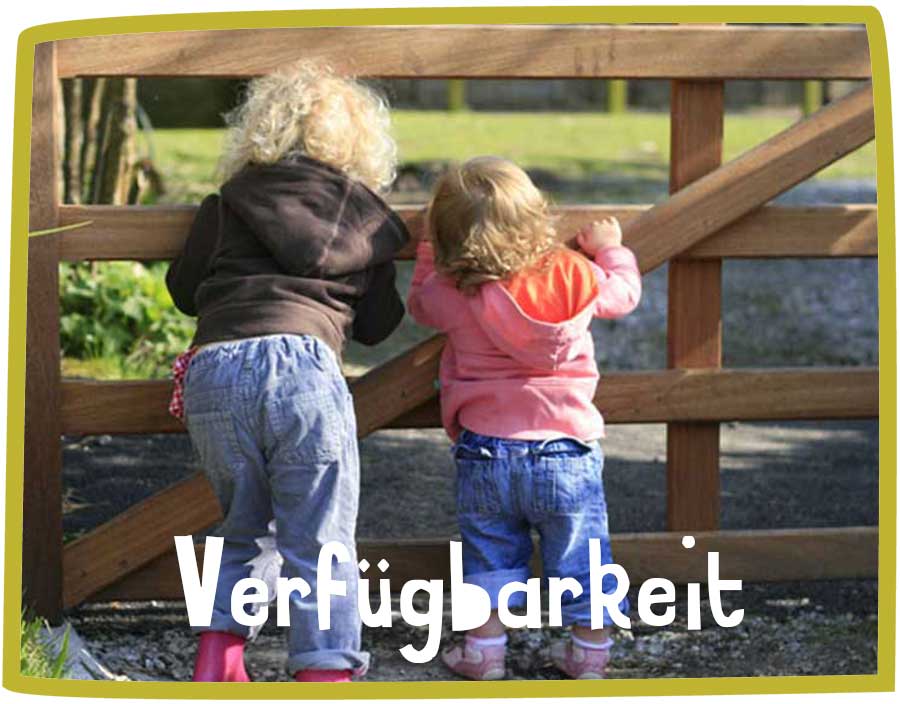 Two children in wellies peering through a brown wooden gate.