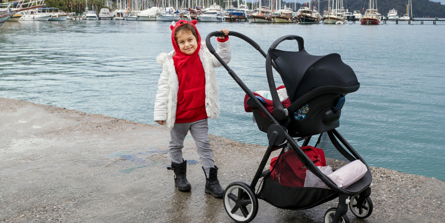 young girl pushing pram on water front with boats in back ground