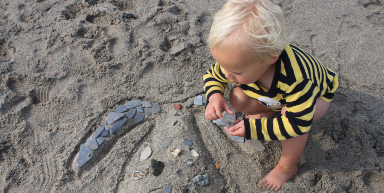 camera looking down on a toddler with blonde hair playing n the beach with pebbles